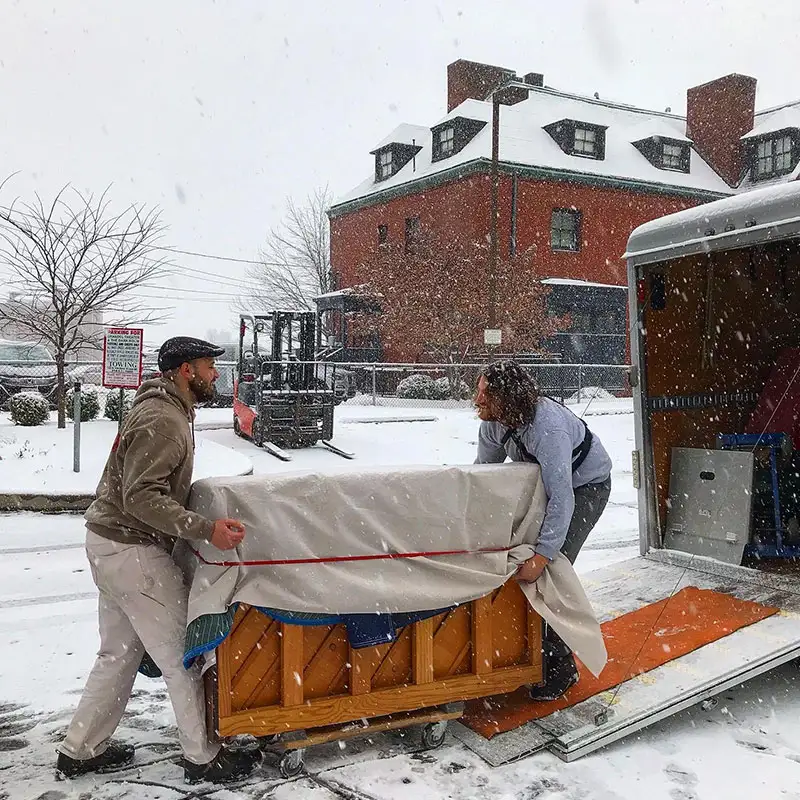 Movers moving a wrapped piano on a dolly through snow
